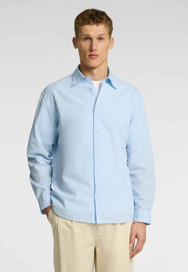 Camisa de manga larga de color azul claro y textura, con cuello abotonado, botones frontales y un corte relajado. Combinada con pantalones de color claro.