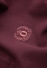 Tela burdeos con tejido texturizado y un emblema bordado que presenta una corona de laurel, una corona, un óvalo y la palabra "ORIGINAL".