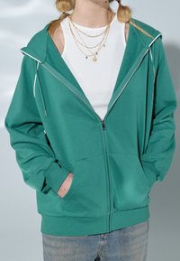 Sudadera verde con cremallera, con una gran capucha, mangas largas, bolsillos frontales y cordones blancos. Lleva encima una camiseta de tirantes blanca con canalé.
