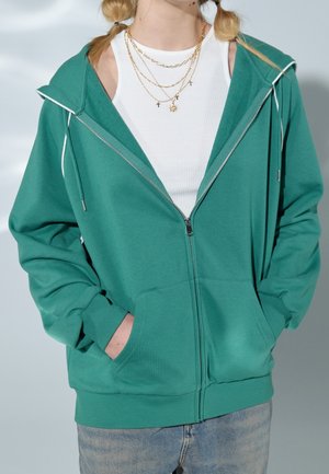 Green zip-up hoodie with a large hood, long sleeves, front pockets, and white drawstrings. Layered over a white ribbed tank top.