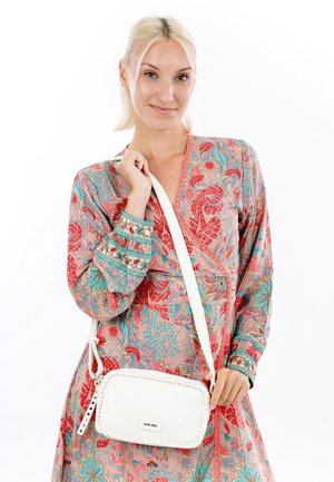 Junge Frau mit blonden Haaren trägt ein buntes Blumenmusterkleid und hält eine weiße Leder-Umhängetasche mit geflochtenem Rand.