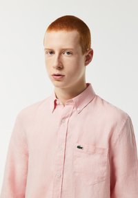 Camicia button-up in lino rosa chiaro con maniche lunghe, una piccola tasca sul petto e un logo verde sulla tasca. Texture liscia e design casual.