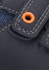 Detalle de zapato de cuero navy con un ojal naranja, costura blanca contrastante y un acento de tela de mezclilla. Presenta un diseño de correa segura.
