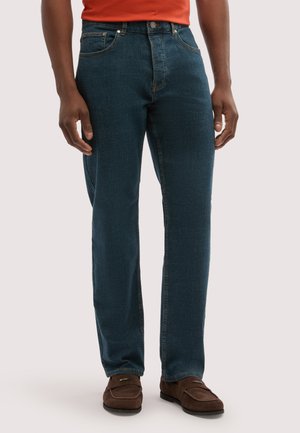 Mann trägt dunkelblaue Jeans mit geradem Bein, braune Loafers und ein oranges Hemd, steht vor einem schlichten hellen Hintergrund.