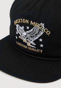 Cappello nero con un'aquila bianca ricamata che tiene un fulmine, circondata da stelle, e testo dorato "BRIXTON MFG CO. CUSTOM QUALITY."