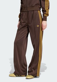 Pantaloni de trening din poliester maro cu dungi laterale galbene, talie elastică și buzunare cu fermoar. Prezintă un mic detaliu cu logo galben.