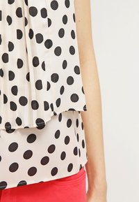 Blusa a pois con un design plissettato, caratterizzata da grandi pois neri su uno sfondo beige chiaro, abbinata a un indumento rosso nella parte inferiore.
