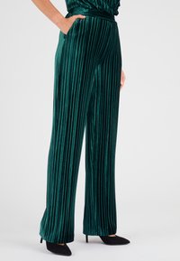 Pantaloni in velluto verde, plissettati, con design a gamba larga e tasche laterali. Accentuati da una cintura liscia e indossati con scarpe nere a punta.