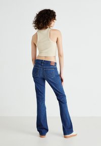Flared denim jeans in dark blue with a classic five-pocket design. A textured tan label on the back waistband and yellow stitching details.