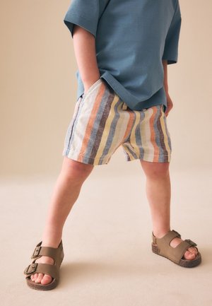 Kind steht mit Händen in den Taschen, trägt ein blaues Hemd, mehrfarbige gestreifte Shorts und braune Sandalen auf hellem Hintergrund.