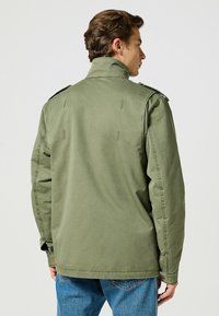 Olivgrön bomullsjacka med ståkrage, axelpartier och synliga sömnadsdetaljer, i kombination med blå denimbyxor.