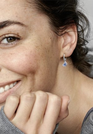 Boucles d'oreilles en argent avec un pendentif en pierre précieuse bleue, présentant des détails complexes et une finition polie, portées par une personne vêtue d'un vêtement gris clair.