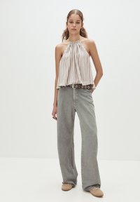 Donna in piedi che indossa un top senza maniche a righe, jeans grigi a gamba larga con una cintura nera con borchie, e scarpe beige su uno sfondo neutro.