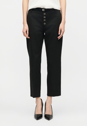 Žena nosi čierne skrátené nohavice s dekoratívnymi gombíkmi a úzkym čiernym opaskom, doplnené čiernymi špicatými vysokými opätkami.