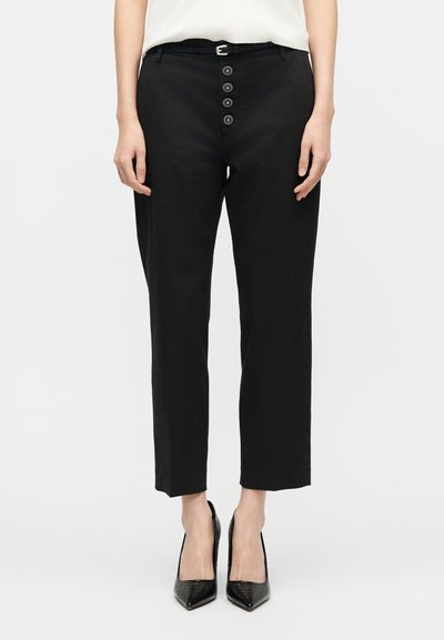 Γυναίκα με μαύρο κομμένο παντελόνι διακοσμημένο με κουμπιά και λεπτή μαύρη ζώνη, συνδυασμένο με μαύρες μυτερές ψηλές γόβες.