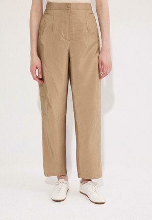 Pantalon beige taille haute à jambes droites avec bouton et fermeture éclair devant, porté avec des chaussures blanches à lacets sur fond uni.