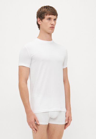 Weißes kurzärmliges T-Shirt aus glattem Material, Rundhalsausschnitt, sitzt eng am Körper. Getragen mit passenden weißen Slips steht das Model vor einem schlichten Hintergrund.