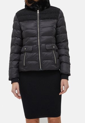 Sort quiltet jakke med pelskrave, lynlås foran, klap og lynlåslommer båret over en sort ribstrikket nederdel.