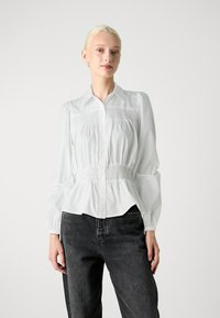 Bluză albă cu nasturi, cu mâneci lungi și voluminoase, detalii cu smocuri la talie și un tiv fluid. Asortată cu blugi închiși la culoare.