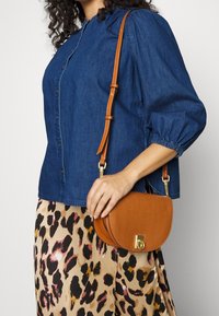 Donna che indossa una camicia di jeans e una gonna a stampa leopardata, mentre tiene in mano una piccola borsa a tracolla in cuoio marrone con chiusura dorata.