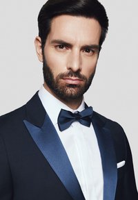 Ciemny garnitur z satynowym kołnierzykiem w kształcie szala, biała koszula i czarna muszka. Krótkie, starannie stylizowane włosy oraz zadbana broda.