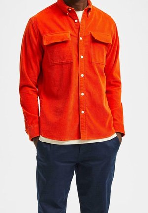 Homme portant une chemise en velours côtelé orange vif à boutons avec poches poitrine et pantalon bleu foncé, mains dans les poches, sur fond blanc uni.
