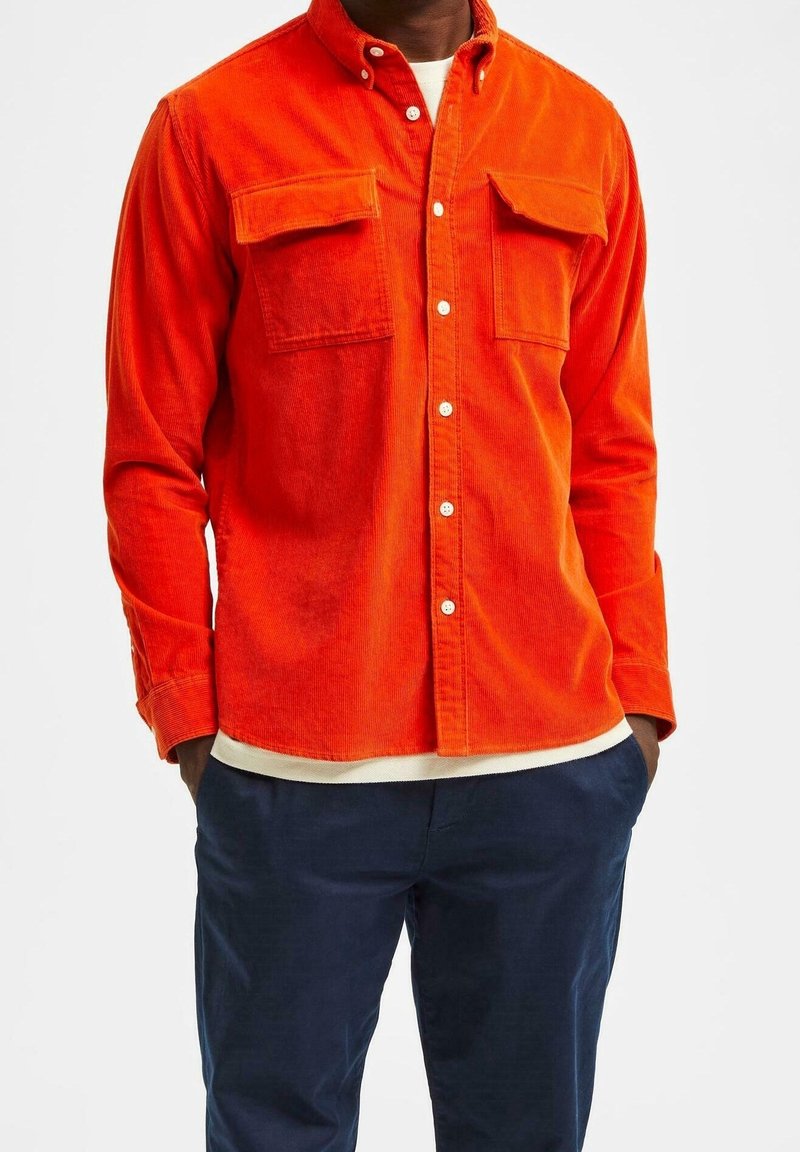 Homme portant une chemise en velours côtelé orange vif à boutons avec poches poitrine et pantalon bleu foncé, mains dans les poches, sur fond blanc uni.