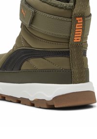 Bota verde oliva con un upper texturizado, acentos negros, entresuela blanca y detalle del logo en naranja. Cuenta con un cuello acolchado y correa ajustable.