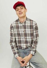 Geruit flanellen shirt in crème, bruin en marineblauw met knoopsluiting; model draagt lichtblauwe spijkerbroek en een rode pet.