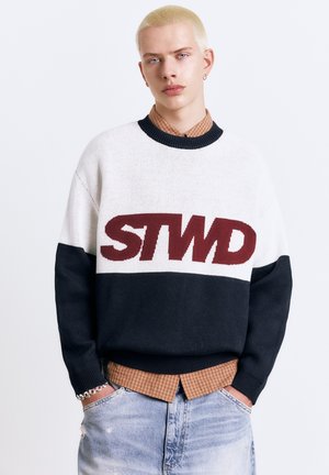 Mladá osoba s krátkymi blond vlasmi, oblečená v bielom a čiernom svetri s nápisom "STWD", hnedou károvanou košeľou a svetlomodrými džínsami, s rukami v vreckách.