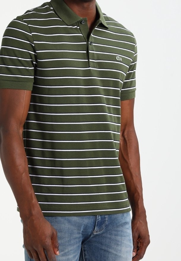 Homme portant un polo vert à manches courtes avec des rayures horizontales blanches et un jean bleu, debout devant un fond uni.