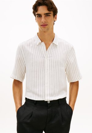 Ung mand iført en hvid kortærmet skjorte med tynde, lodrette striber og sorte bukser, stående med hænderne i lommerne.