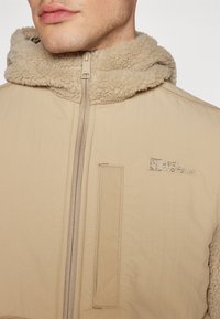 Beige Jacke mit einer weichen, strukturierten Kapuze. Verfügt über einen Reißverschluss, ein kleines Logo auf der Brust und eine aufgesetzte Tasche auf der Vorderseite.