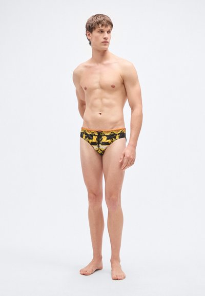 Μαγιό σλιπ σε μαύρο με κίτρινο σχέδιο που απεικονίζει άλογα, με ελαστική μέση και ελάχιστη κάλυψη. Το υλικό φαίνεται λείο και εφαρμοστό.