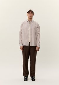 Mann iført beige stripet skjorte med knapper, hvit t-skjorte, mørkebrune bukser, svarte sko og beige baseballcaps, stående mot ensfarget bakgrunn.