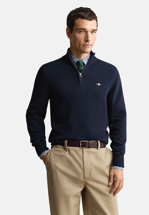 Navy zip-neck sweater with ribbed cuffs and collar, worn over a striped dress shirt, paired with khaki pants and a brown leather belt.