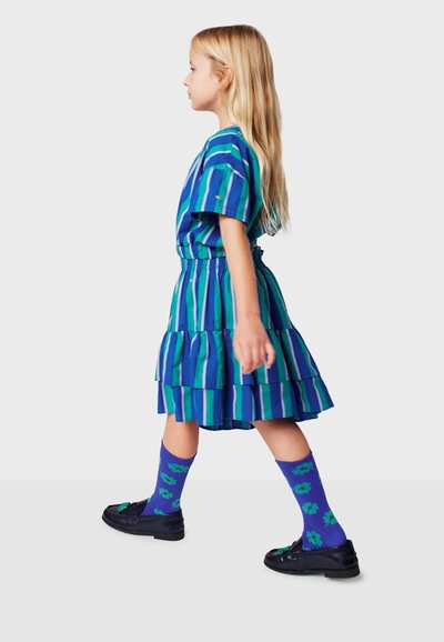 Abito a righe blu e verdi con balze a strati, indossato con calze viola con motivi floreali e scarpe nere.