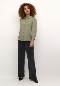 Camicia verde oliva con colletto e maniche lunghe a sbuffo; abbinata a jeans neri a vita alta con gamba larga e scarpe nere a punta.