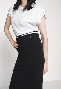 Vit kortärmad t-shirt kombinerad med en figurnära svart kjol med resårmidja och CK-logotyp på framsidan. Slät tygstruktur.