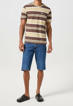 Mies, jolla on päällään ruskean ja beigen raidallinen t-paita, siniset farkkushortsit ja mustat slip-on-kengät, seisoo yksinkertaisen vaalean taustan edessä.