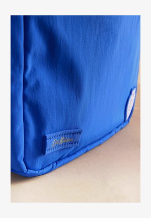 Hjørne af en lys blå stofpose med Cath Kidston-logo i guldtryk syet på en beige overflade.