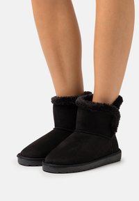 Un par de botas negras hasta el tobillo con forro de piel de peluche, llevadas sobre piernas desnudas contra un fondo claro y liso.