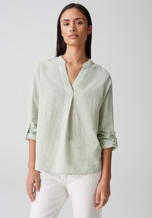 Blusa di lino verde chiaro con scollo a V e maniche arrotolate, caratterizzata da una vestibilità rilassata e una leggera trama. Indossata con pantaloni bianchi.