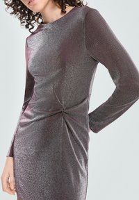 Kobieta ubrana w błyszczącą, dopasowaną sukienkę z długim rękawem i marszczeniem w talii, na jednolitym tle.