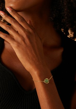 Een persoon met een donkere huid draagt een zwarte top en een gouden armband met een ronde groene steen, terwijl hun hand voorzichtig hun nek aanraakt.