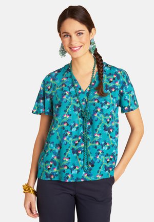 Frau mit geflochtenem Haar, die eine türkise Bluse mit Blumenmuster, lange Perlenkette, große Ohrringe, goldene Armreifen und dunkle Hosen trägt, lächelnd mit der Hand in der Tasche.