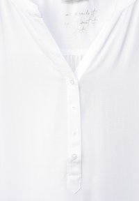 Camicia bianca con scollatura a V, chiusura con bottoni e dettagli plissettati sul collo. Il tessuto sembra liscio e leggero.