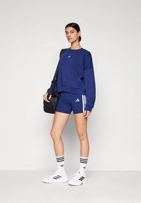 Nő kék Adidas pulóverben és rövidnadrágban, fehér Adidas zoknival és tornacipővel, fekete válltáska a kezében, sima világos háttér előtt.