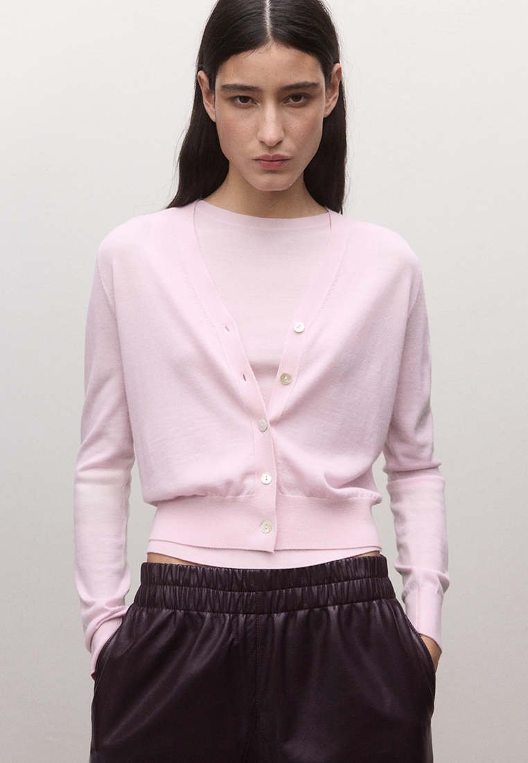 Femme aux longs cheveux foncés porte un cardigan rose clair boutonné et un haut assorti, associé à un pantalon noir ample à taille élastique. Mains dans les poches.