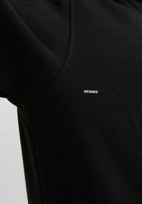 Schwarzes Baumwoll-T-Shirt mit rundem Halsausschnitt. Beinhaltet kleinen weißen Text "DISTANCE" auf der linken Schulter. Weicher Stoff, minimales Design.